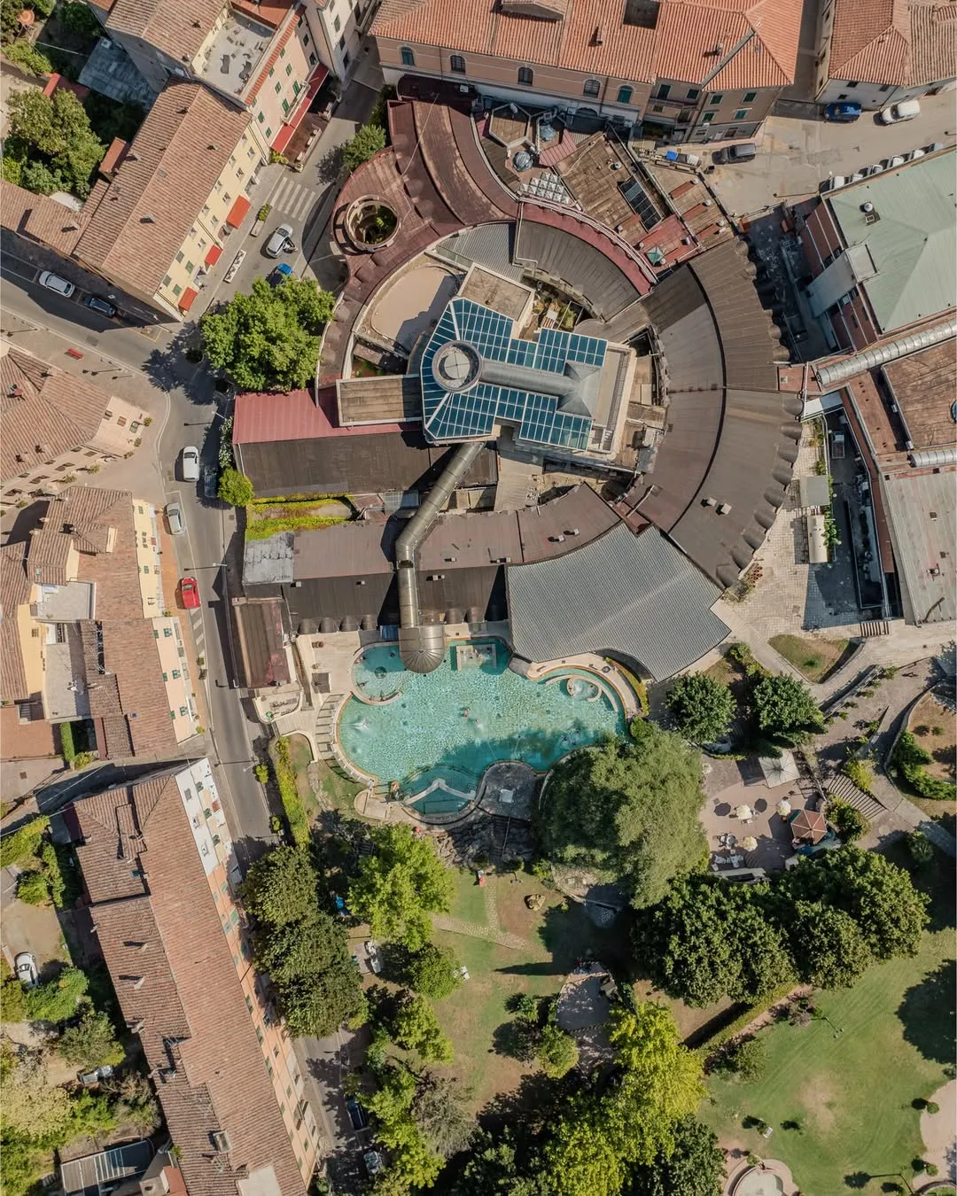An overhead drone shot showcasing a bustling town square with a prominent spa complex featuring multiple pools, surrounded by traditional buildings and green spaces.