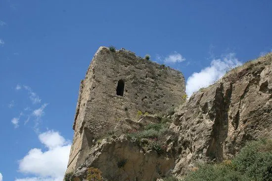 An ancient stone tower or fortress ruin stands prominently on a rocky hill under a bright blue sky with scattered clouds.