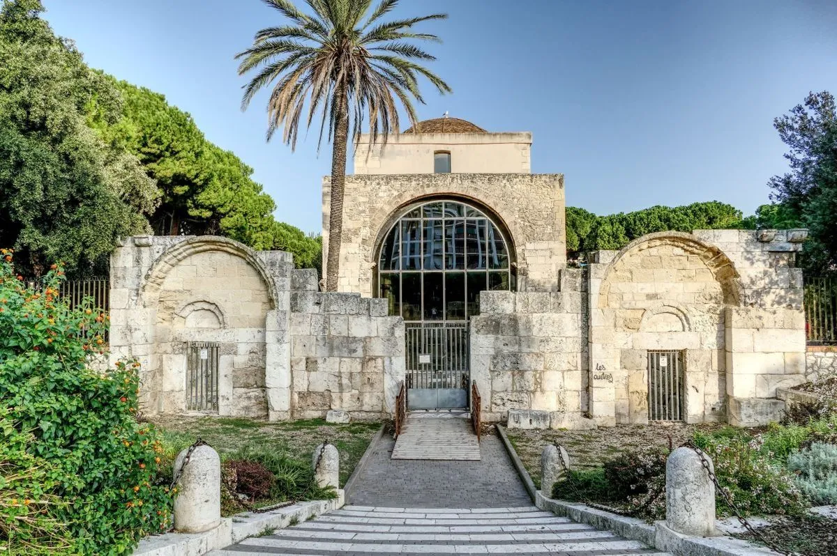 The image prominently features a historic stone building with a large arched window, flanked by stone walls and lush greenery under a clear blue sky. A tall palm tree stands beside the building. A paved path and steps lead towards the building's entrance.