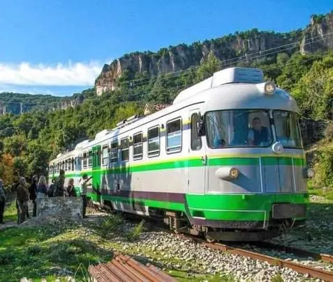 An outdoor shot of a green and white train on tracks, with lush green hills and rocky mountains under a bright blue sky. There are some people standing near the train.