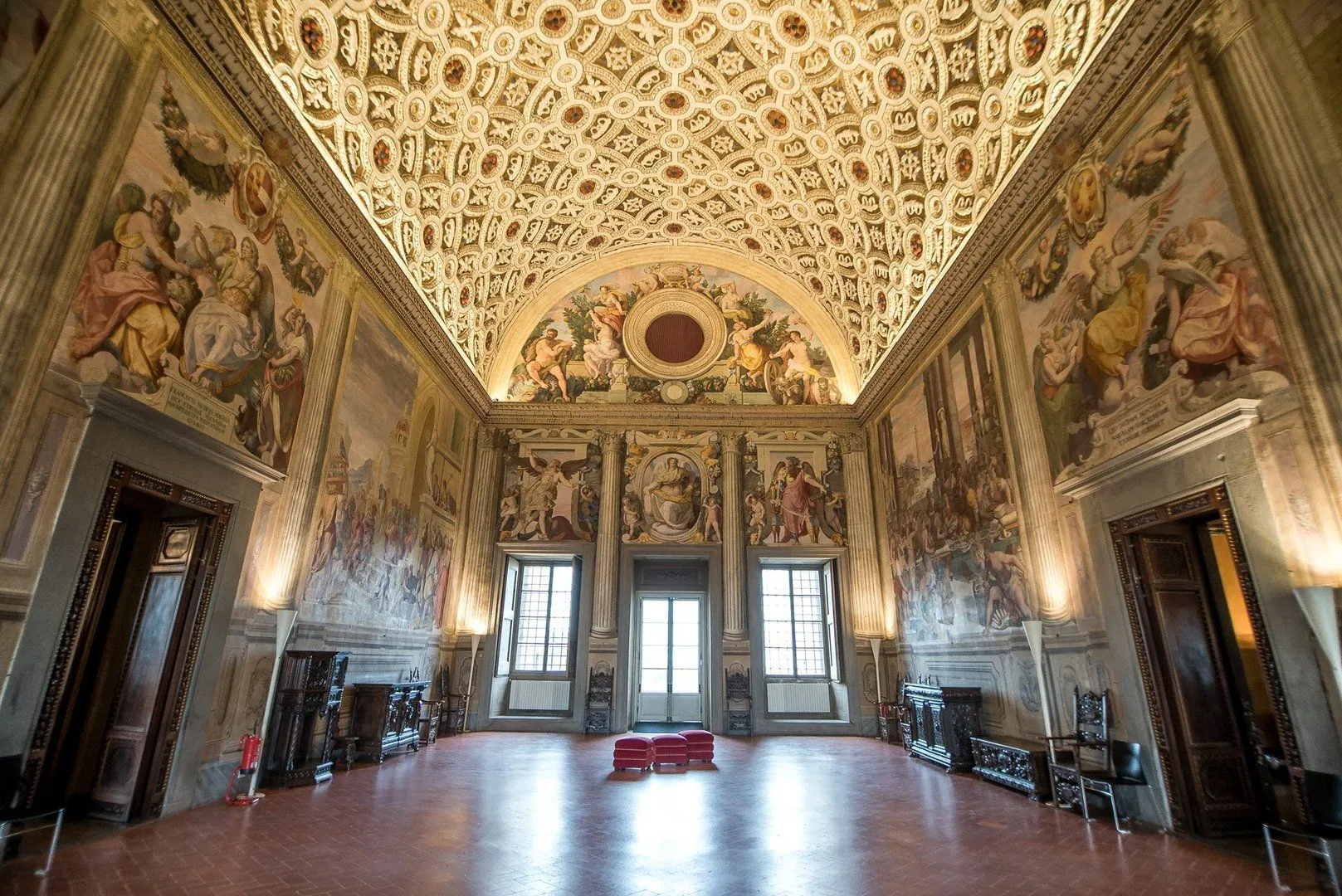 A grand hall featuring an intricately decorated ceiling and walls adorned with large-scale frescoes. The room is well-composed, highlighting the ornate details of the historical interior. The lighting effectively illuminates the upper sections, capturing the richness of the artwork.