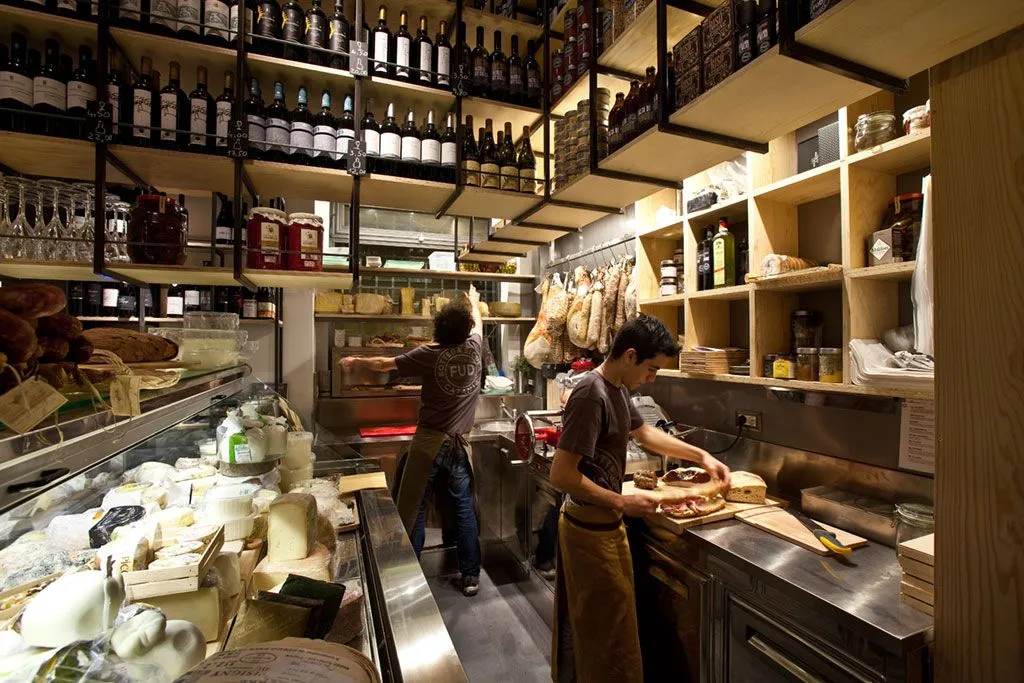 An interior shot of a bustling deli or specialty food shop, showcasing shelves stocked with wine and other products, a cheese display, and two workers attending to tasks behind the counter, one preparing food.