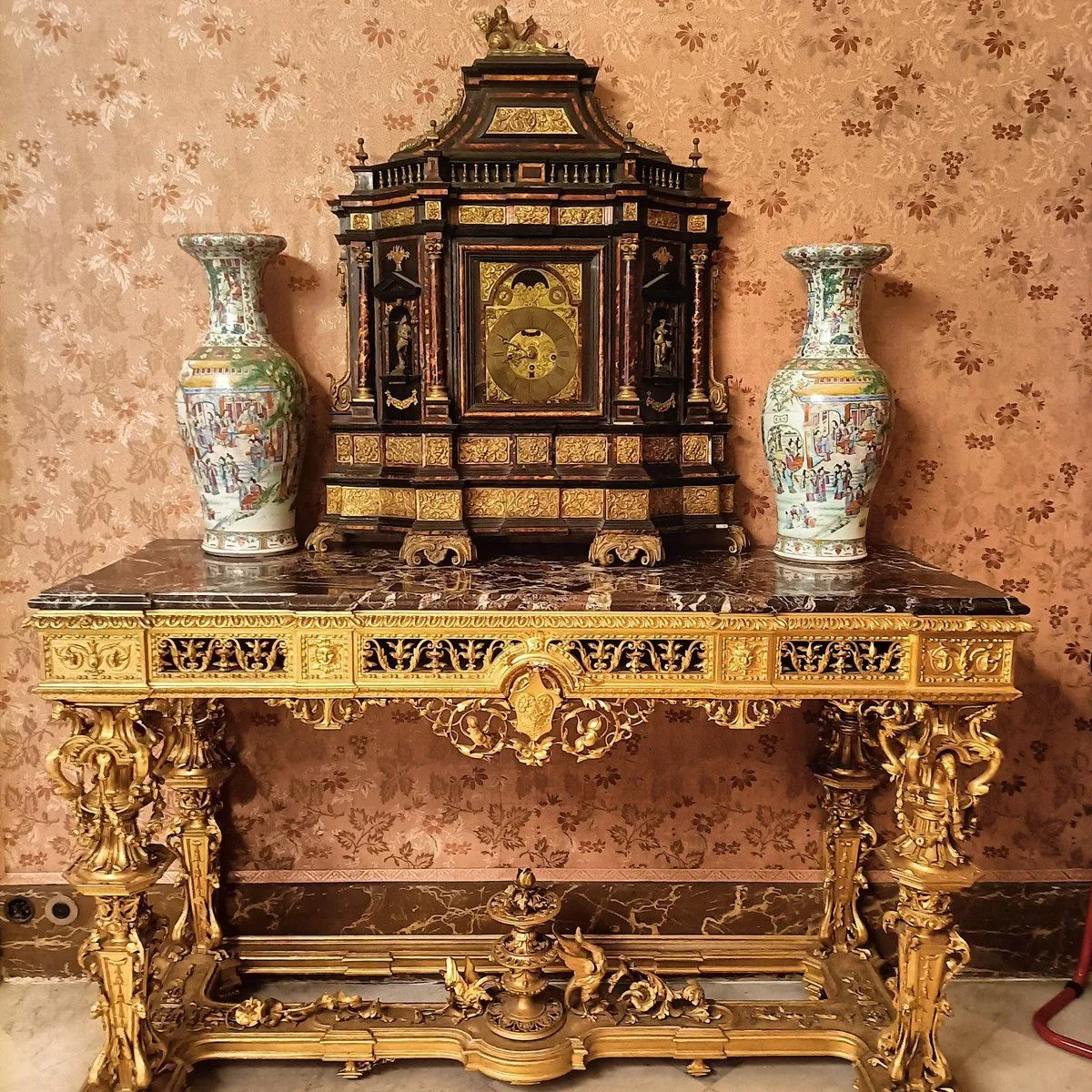 A detailed image showcasing an antique ornate golden table with a marble top, flanked by two decorative vases, and a grand clock centerpiece, set against a classic patterned wallpaper.