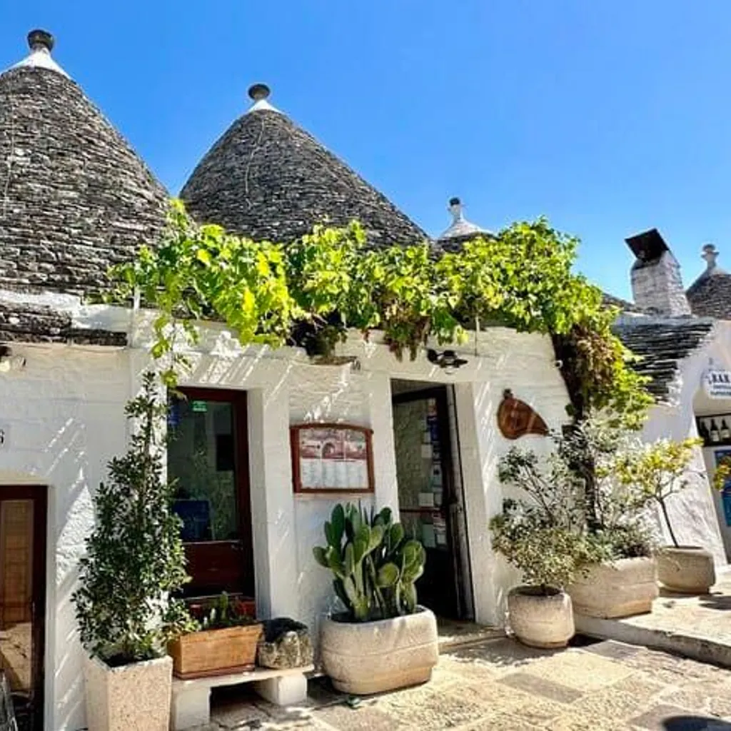 An editorial style image showcasing the unique conical-roofed Trulli houses, characteristic of Puglia, Italy. The image is well-composed, highlighting the architectural details and local flora under a clear blue sky.