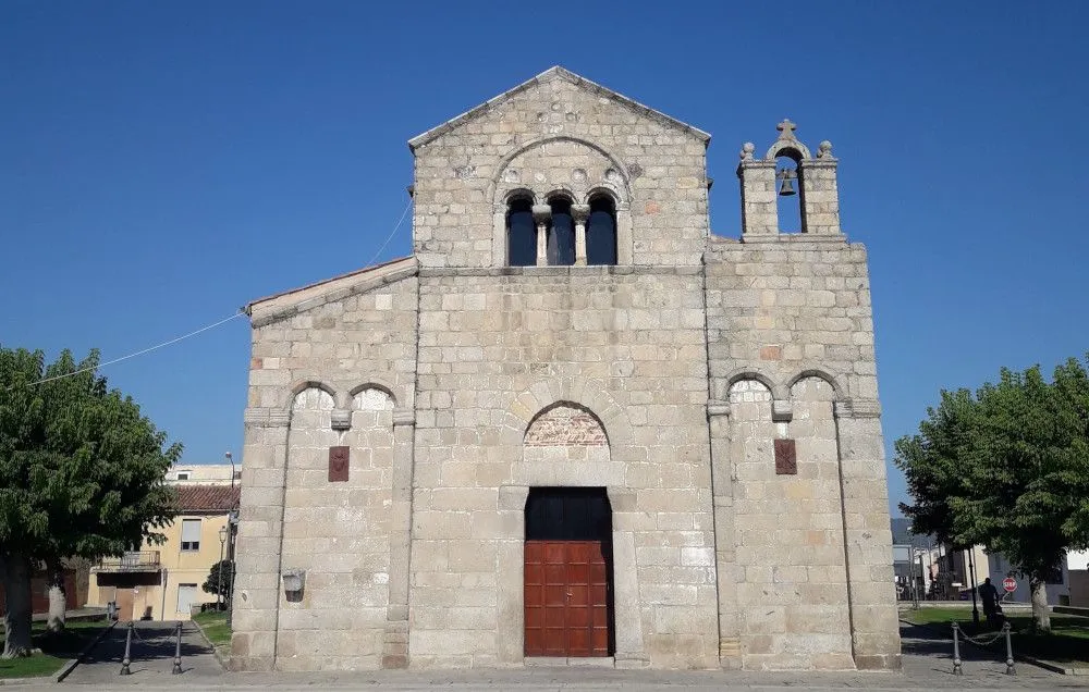 A well-lit, frontal view of an ancient stone church under a clear blue sky, showcasing its rustic stone facade, arched windows, and small bell tower.