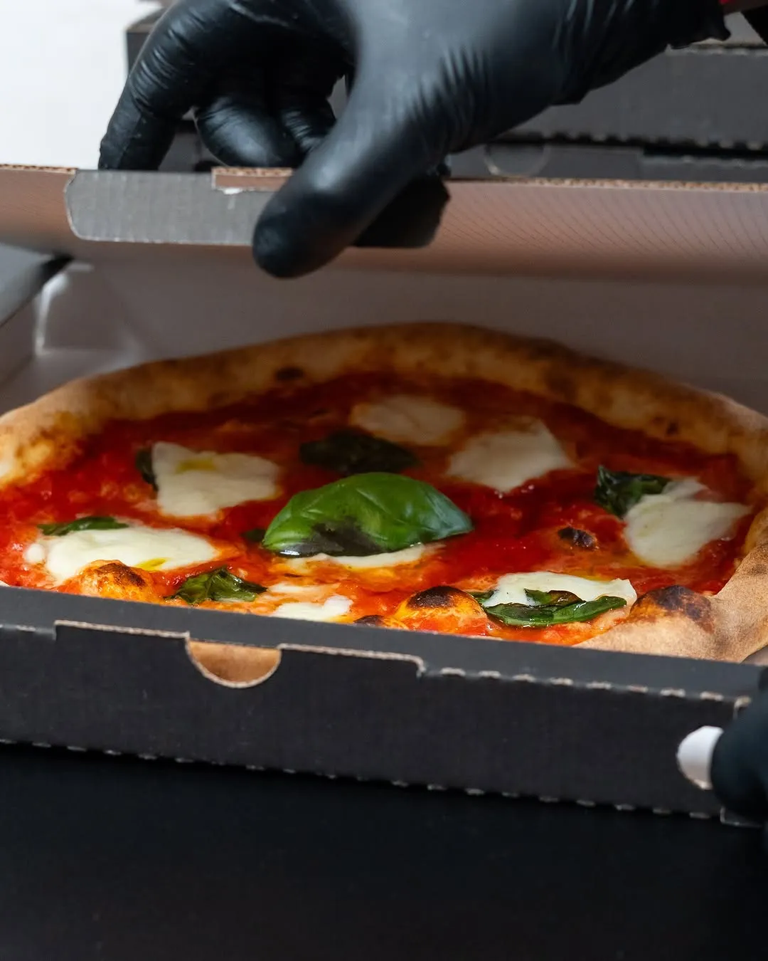 A top-down shot of a freshly baked pizza in an open pizza box, with hands in black gloves holding the lid. The pizza has a classic Margherita style with vibrant red sauce, white mozzarella, and fresh green basil leaves.