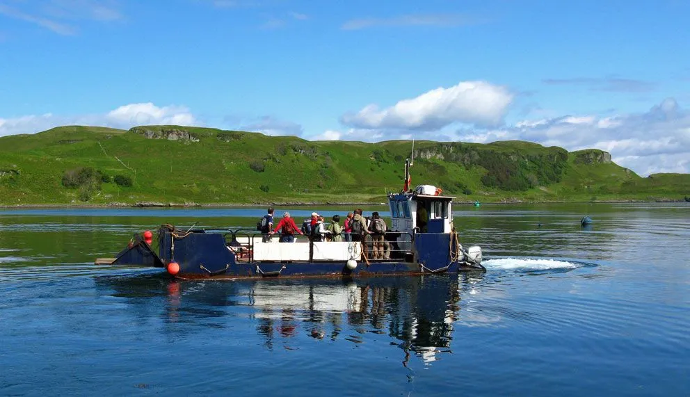 A small boat carrying several passengers traverses a body of blue water, with verdant green hills rising in the background under a clear sky with scattered white clouds.