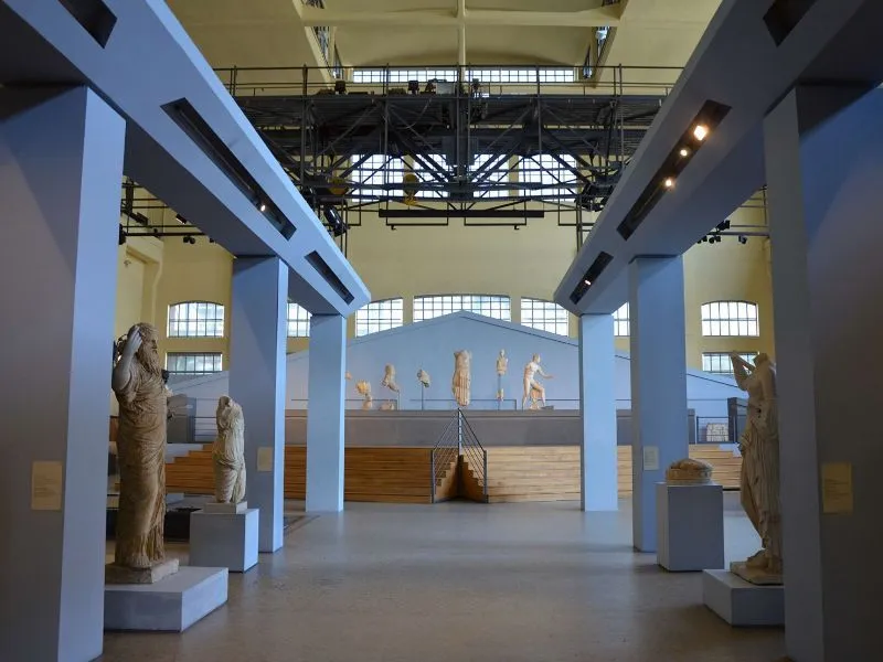 An interior view of a museum or exhibition hall showcasing classical statues. The space is large with high ceilings, featuring architectural beams and large windows in the background. The statues are arranged on platforms and pedestals, with a central tiered display for several smaller sculptures.