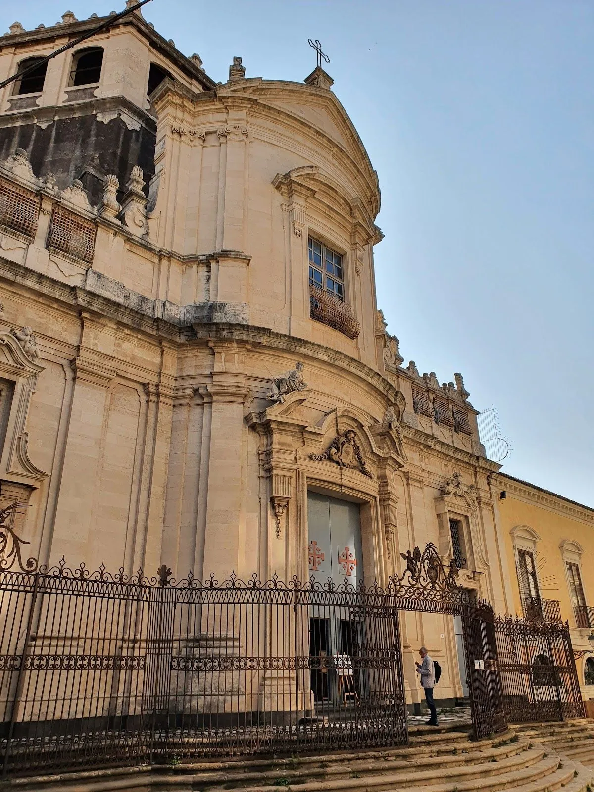 An impressive, ornate stone building with a large entrance and a decorative iron fence in the foreground. The architecture suggests an historical or grand structure, possibly a church or palace. The image is captured under clear skies, with natural lighting highlighting the details of the facade.