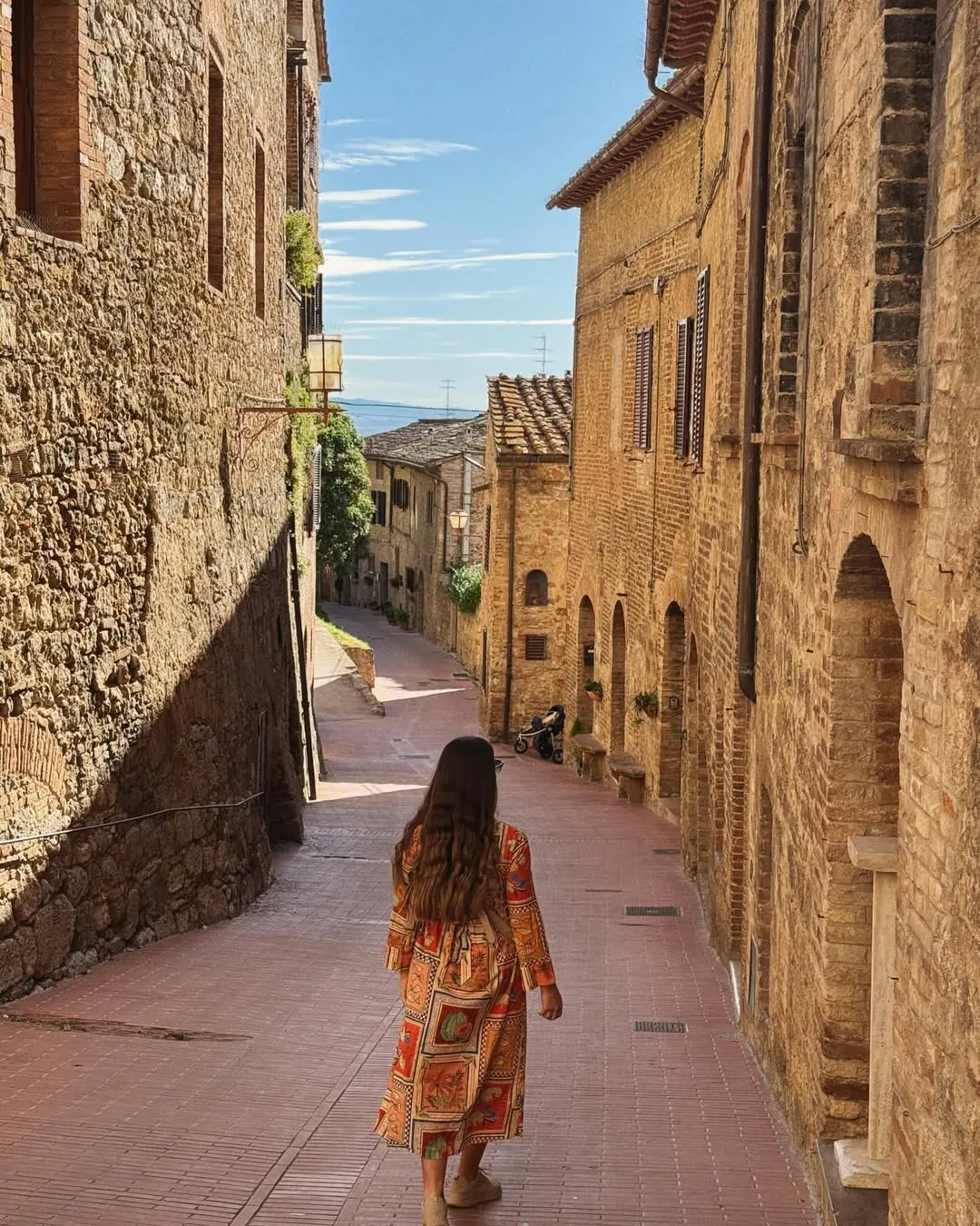 An outdoor photo of a person walking down a narrow, sunlit stone alleyway lined with historic buildings, characteristic of an old European town.
