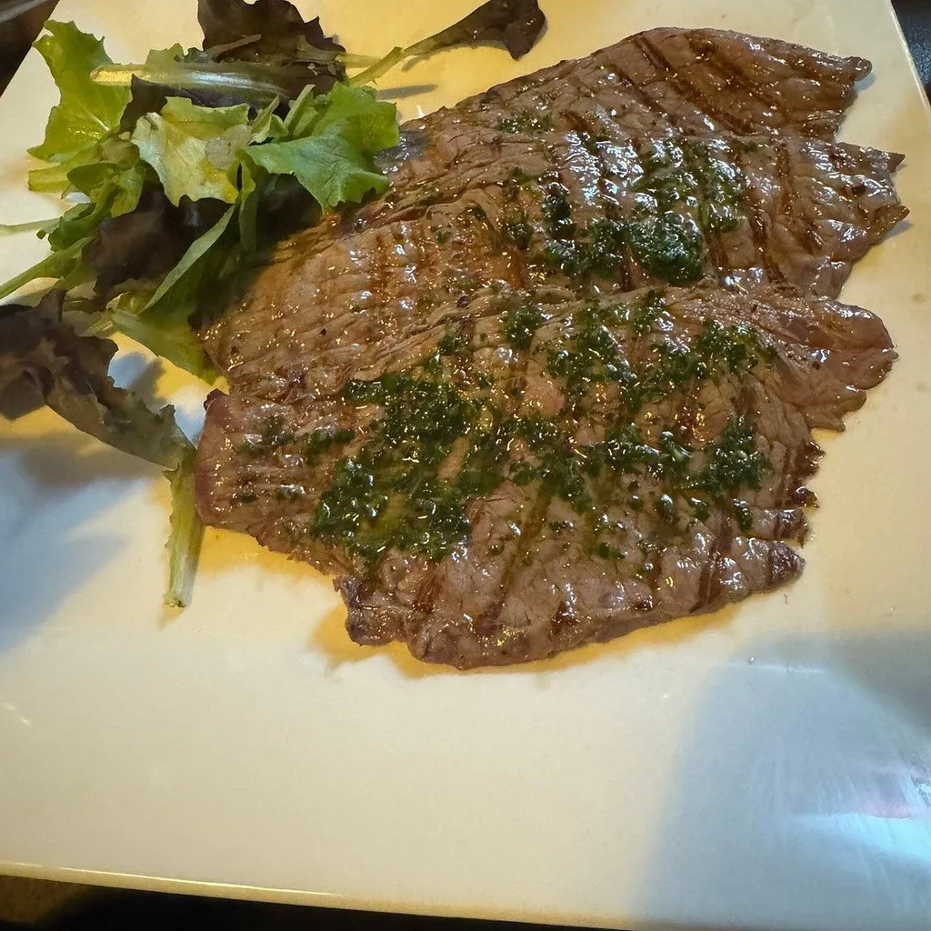 The image features a close-up of a grilled steak with a vibrant green sauce and a side of mixed greens. The key elements, including the steak and sauce, are sharp and in focus, contributing to a strong photo-realistic appearance. The composition is effective, drawing attention to the main dish. However, the lighting is somewhat uneven, leading to some glare on the steak and less vibrant greens, which could be improved for better overall illumination and color accuracy.