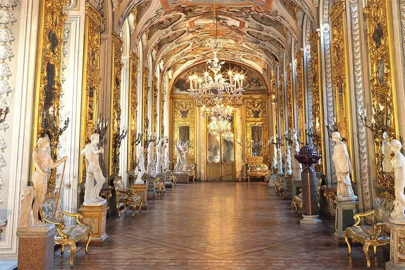 An opulent and grand interior hallway or gallery, richly decorated with gold leaf, intricate frescoes on the vaulted ceiling, and classical statues lining the sides. Large, sparkling chandeliers illuminate the expansive space. The floor appears to be polished wood or parquet, reflecting some of the light.