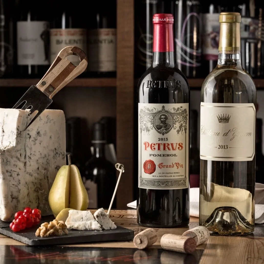 A well-composed still life featuring two prominent wine bottles, one red and one white, arranged alongside a selection of gourmet cheeses, a pear, redcurrants, and walnuts on a dark serving board. The background features a blurred wine rack, providing context and depth.