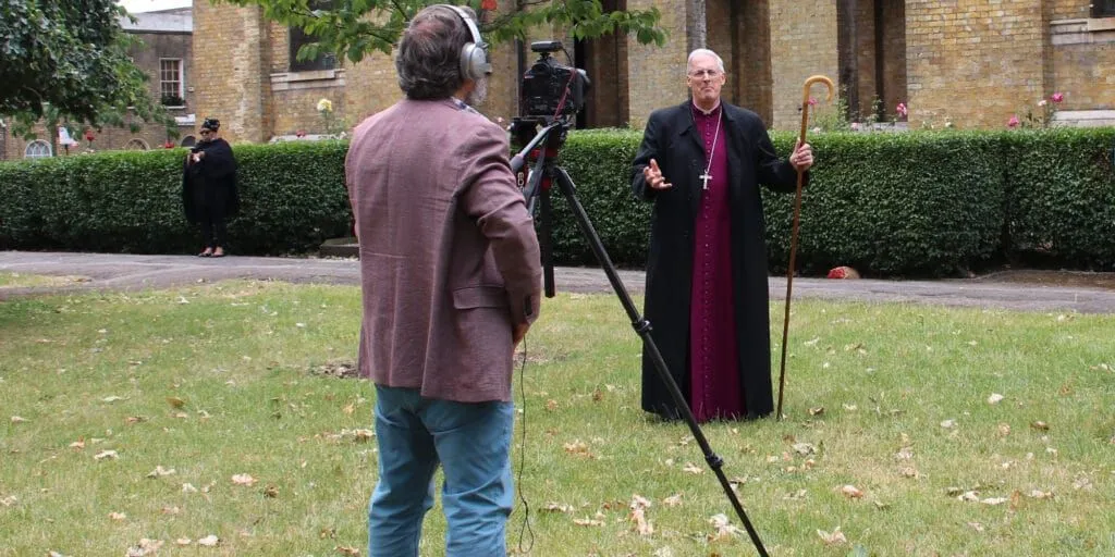 The image captures an outdoor filming session, with a camera operator in the foreground facing away from the viewer, recording a bishop who is speaking. The scene is well-focused with good color accuracy, and the key subjects are sharp. The lighting is even but somewhat flat, leading to a less dynamic appearance. While the composition clearly presents the interaction, the back of the camera operator dominates the foreground, slightly obscuring the overall view. There are no unwanted elements such as watermarks or promotional graphics.
