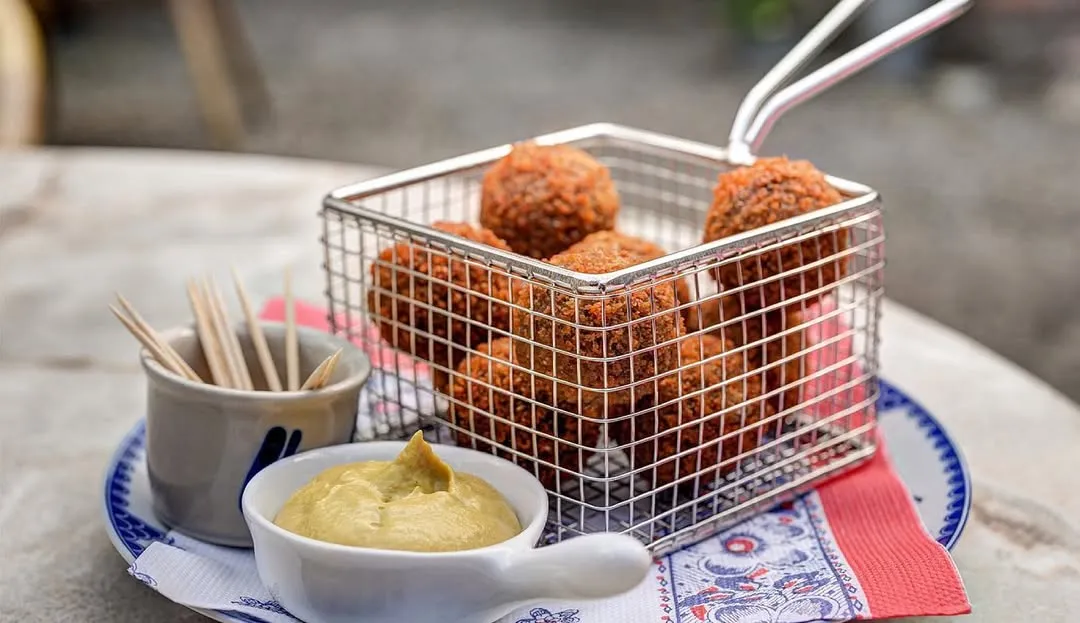 A close-up, well-composed image of fried snacks (likely bitterballen) served in a metal wire basket alongside a bowl of mustard and toothpicks. The lighting is natural and even, highlighting the crispy texture of the food. The colors are vibrant and accurate, making the dish look appealing. The depth of field is shallow, effectively bringing the main subjects into sharp focus while blurring the background.