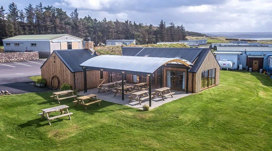 An exterior shot of a modern wooden building with a large outdoor patio area furnished with multiple picnic tables, all under a clear arched canopy. The building features dark roofs and wooden siding, set against a backdrop of green grassy hills and a forested area under a cloudy sky.