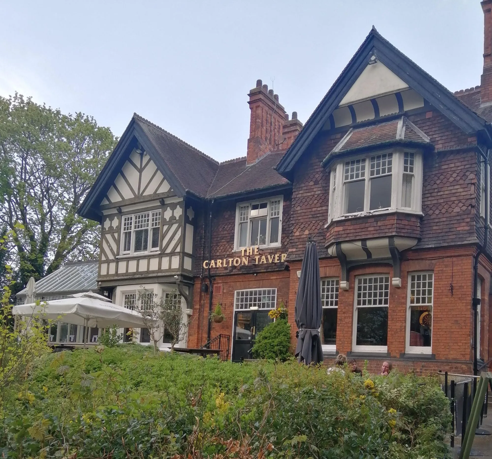 An exterior shot of 'The Carlton Taver' building, showcasing its traditional brick and timber-framed architecture. The image includes outdoor seating with white umbrellas and lush green bushes in the foreground under an overcast sky.