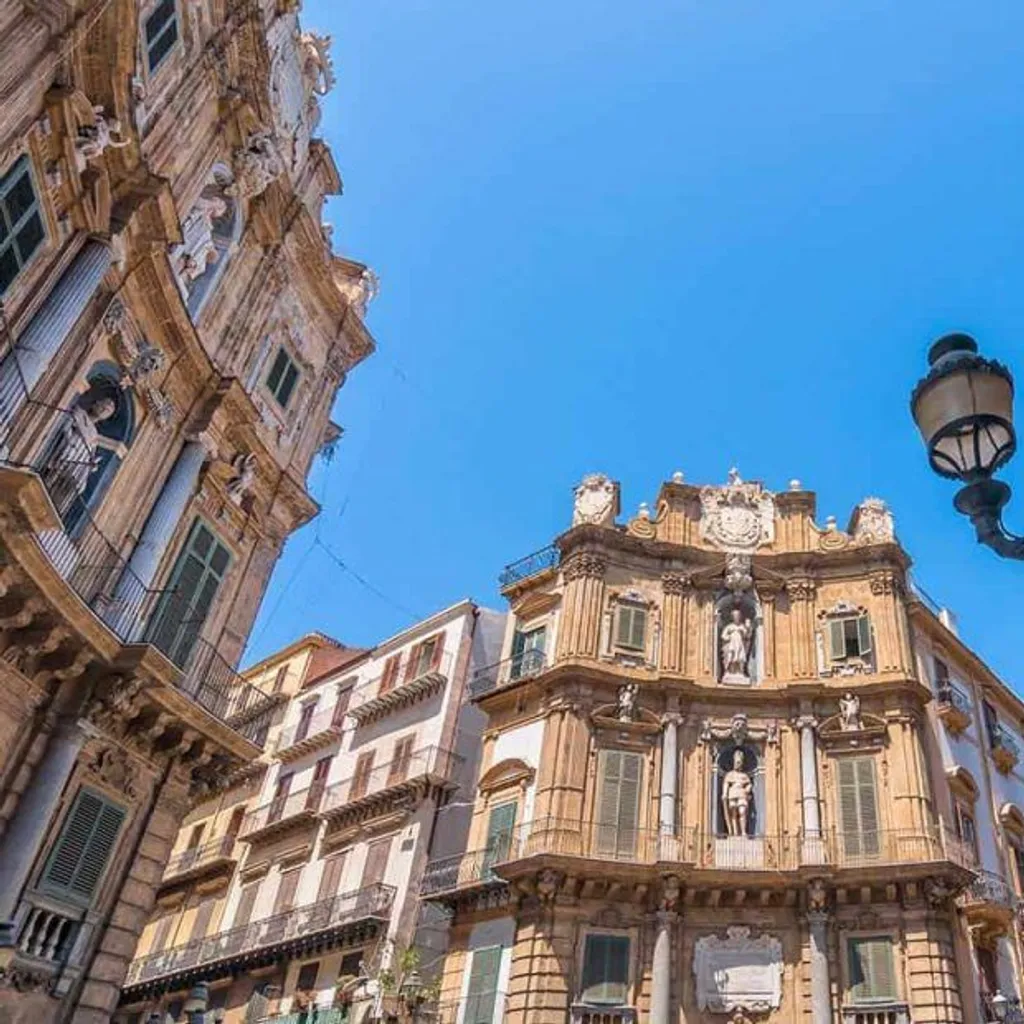 An eye-level shot of ornate historical architecture against a clear blue sky, featuring a prominent, detailed building with statues and balconies, flanked by another building and a decorative street lamp.