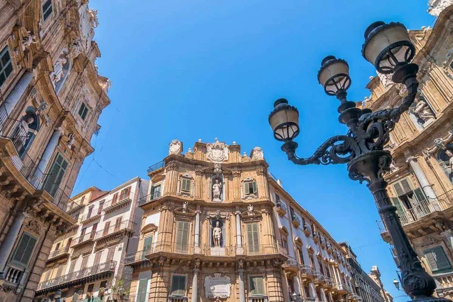 An eye-level shot of ornate historical architecture against a clear blue sky, featuring a prominent, detailed building with statues and balconies, flanked by another building and a decorative street lamp.