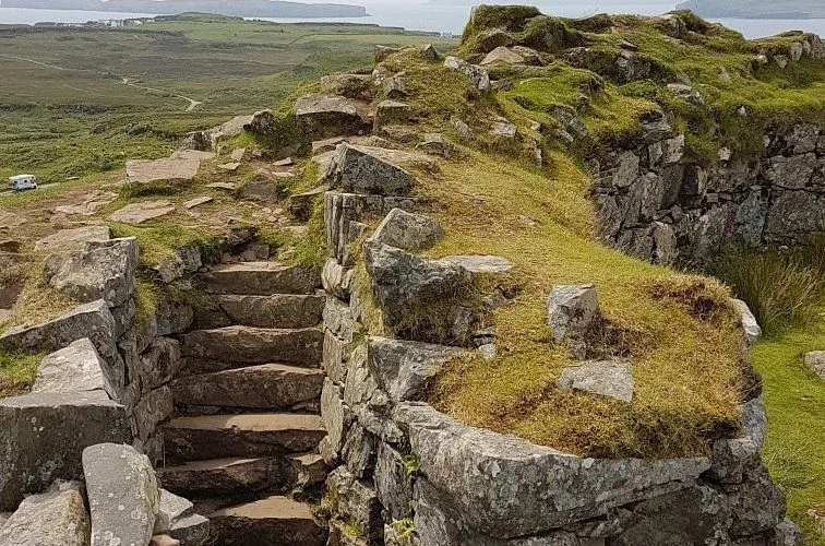 The image is a well-composed and photo-realistic capture of a rocky, grassy landscape with stone steps leading upwards. The key elements in the foreground, such as the steps and rocks, are sharp and in focus. The lighting is natural and even, characteristic of an overcast day, which provides a softer quality to the scene. Colors are accurate, depicting natural greens and greys of the terrain. The composition effectively leads the viewer's eye along the path, with a distant body of water and land visible in the background.