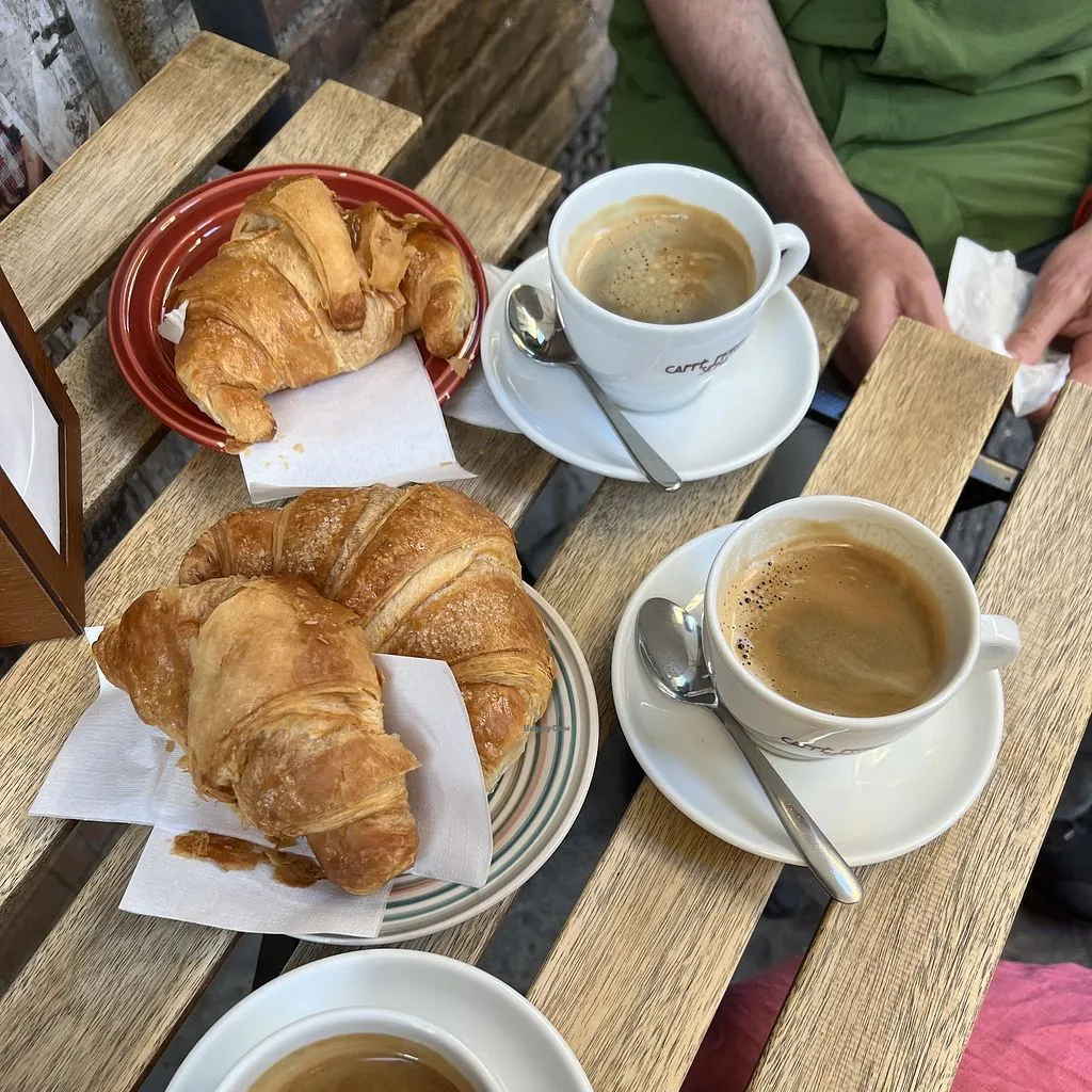 An overhead shot of a breakfast setting featuring three cups of coffee and several croissants on a rustic wooden table, with hands subtly visible in the background.