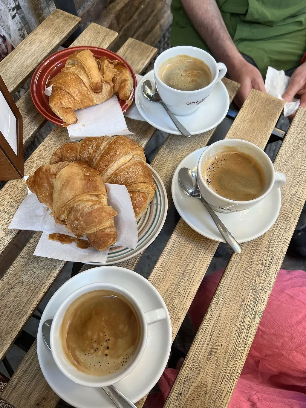 An overhead shot of a breakfast setting featuring three cups of coffee and several croissants on a rustic wooden table, with hands subtly visible in the background.