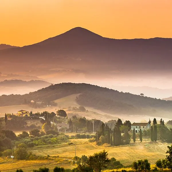 A beautiful panoramic landscape of rolling hills and valleys covered in mist, illuminated by the warm, golden light of sunrise or sunset, featuring scattered traditional houses.
