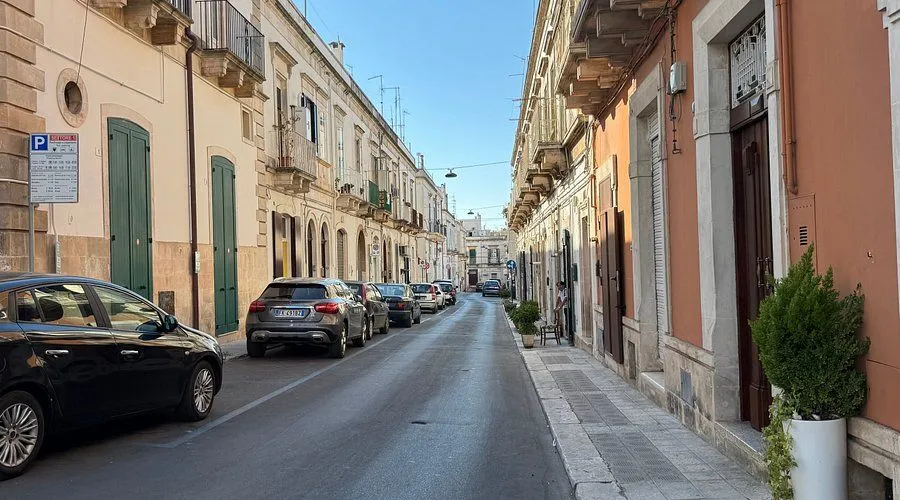 An editorial style image depicting a narrow urban street lined with traditional European-style buildings, with parked cars on one side under a clear blue sky.