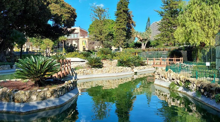 An image depicting a serene park scene with a body of water, lush green plants, and trees under a clear blue sky. A building is visible in the background, adding to the urban landscape.