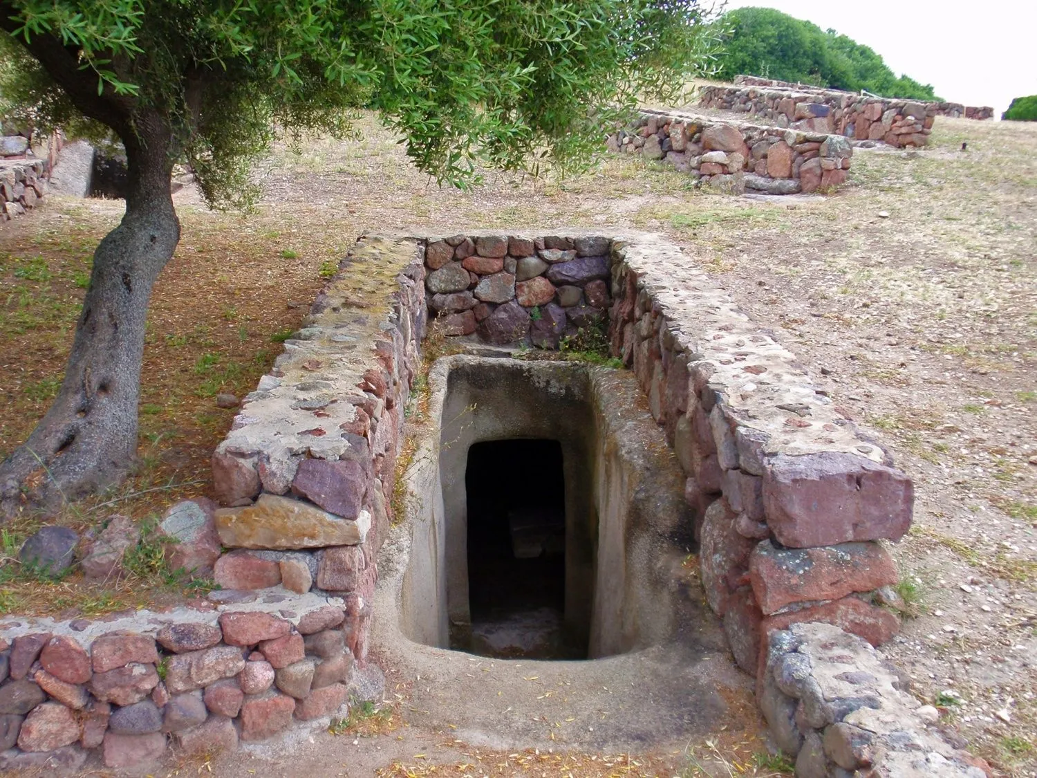 An outdoor photograph capturing the entrance to an ancient or historical underground structure, possibly a tomb or dwelling. The entrance is framed by thick walls constructed from large, reddish-brown stones, leading into a dark, cavernous interior. The structure is set into dry, sparse ground, with a tree partially visible on the left and remnants of other stone walls or terraces in the background under a natural sky.