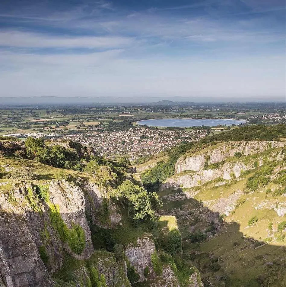 An aerial landscape photograph featuring a deep valley or gorge with rocky cliffs and vegetation in the foreground, leading to a densely populated town and a large lake/reservoir in the mid-distance, all under a clear blue sky with scattered clouds.