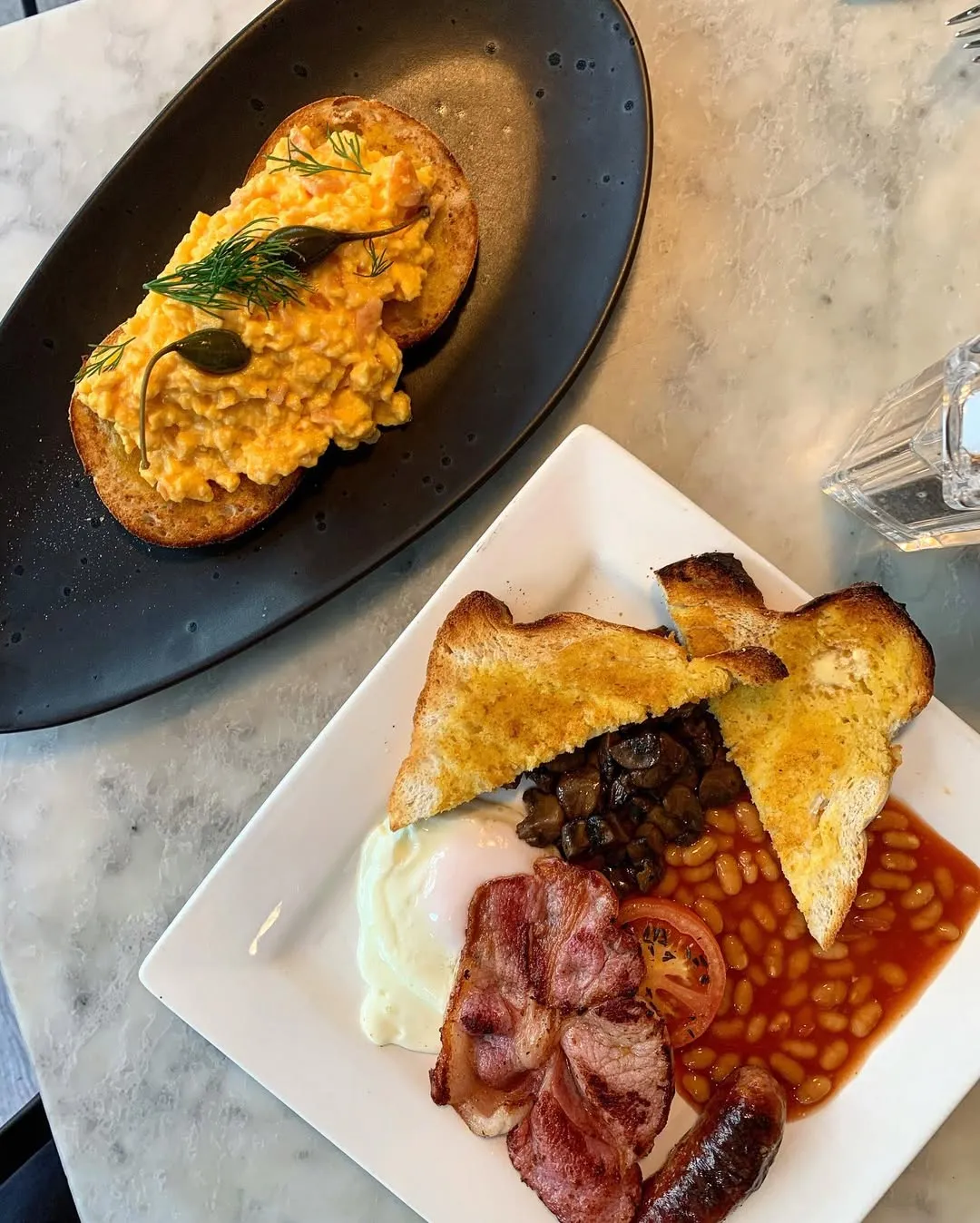The image features two appealing breakfast dishes: scrambled eggs on toast with dill and capers on a black plate, and a full English breakfast on a white plate, including a fried egg, bacon, mushrooms, baked beans, and toast. The lighting is good, colors are accurate, and the main elements are sharp and in focus. The composition is well-balanced for a top-down food shot.
