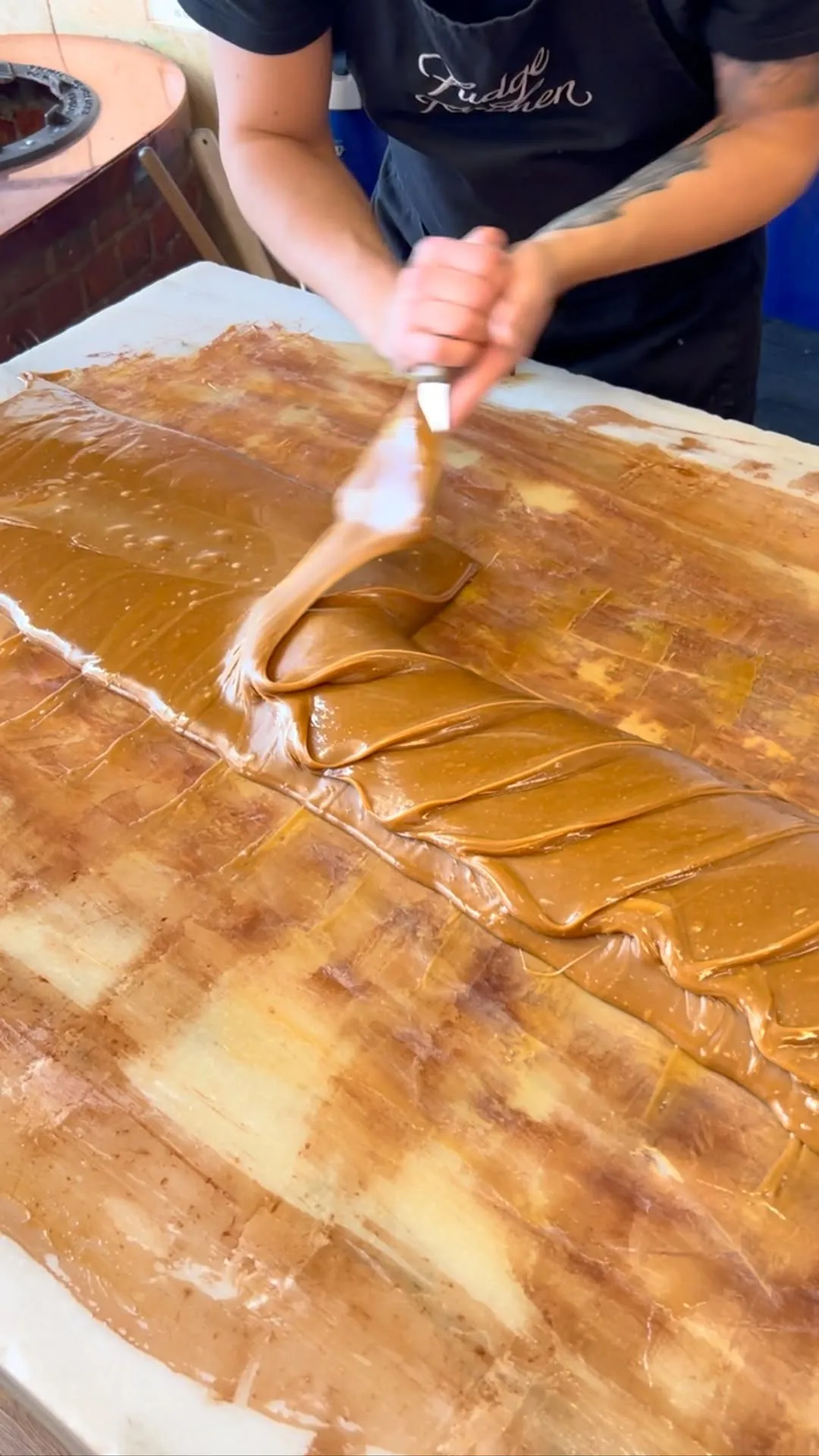A close-up shot of a hand spreading glossy, golden-brown caramel or toffee mixture with a spatula on a flat surface, illustrating a step in confectionery making.