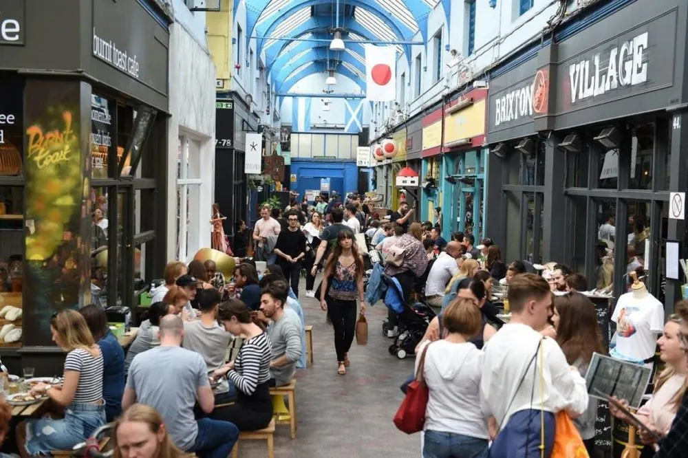 An editorial-style image capturing a bustling indoor market or arcade filled with people. Patrons are seated at tables dining, while others walk through the narrow passage lined with shops and food stalls. The scene is characterized by a distinctive blue arched ceiling.