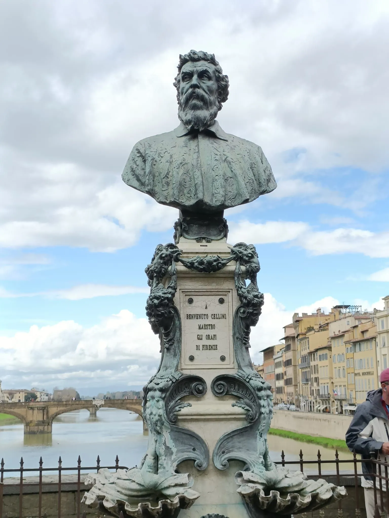 A bronze bust of Benvenuto Cellini on a decorative pedestal, set against the backdrop of the Arno River, Ponte Vecchio, and historic buildings in Florence, Italy, under a partly cloudy sky.