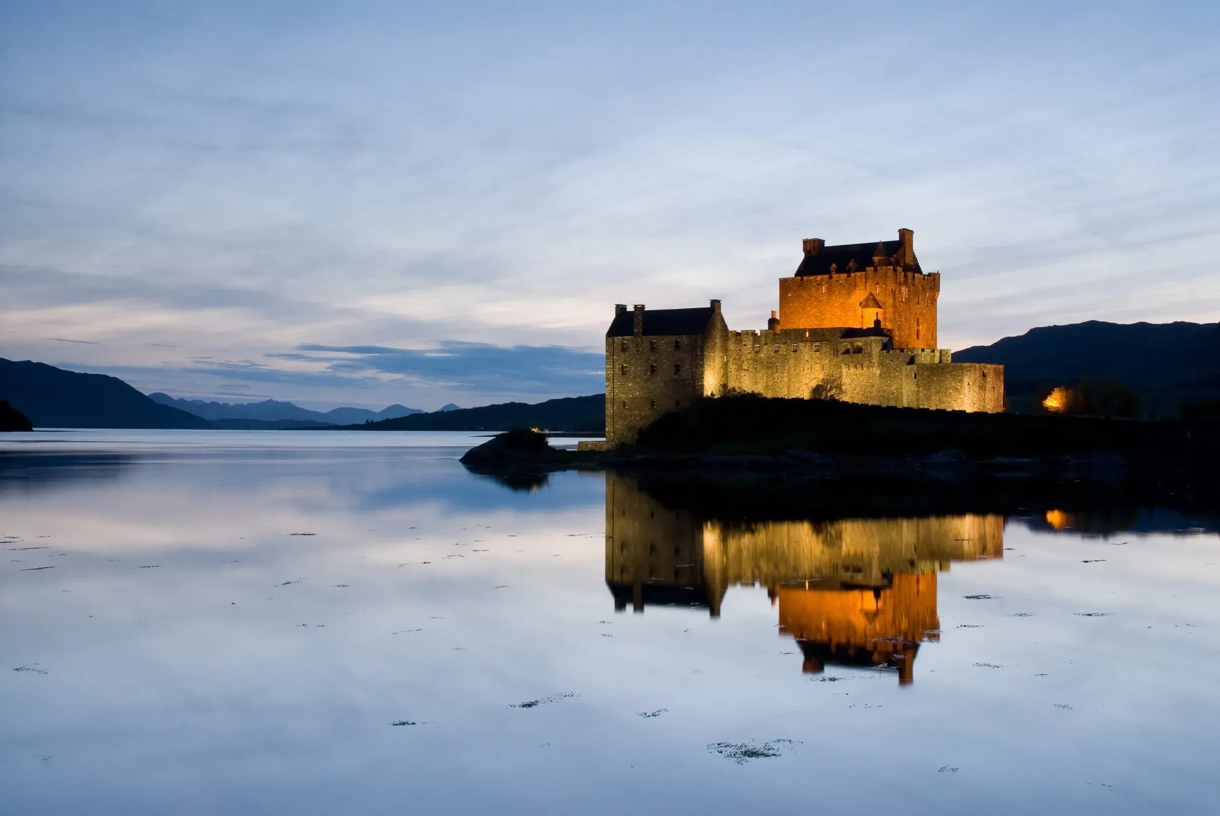 An exceptionally well-composed and visually striking photograph of a castle illuminated at twilight, reflected in still water. The image is sharp, photo-realistic, and beautifully lit, showcasing the architectural details of the castle and the serene environment.