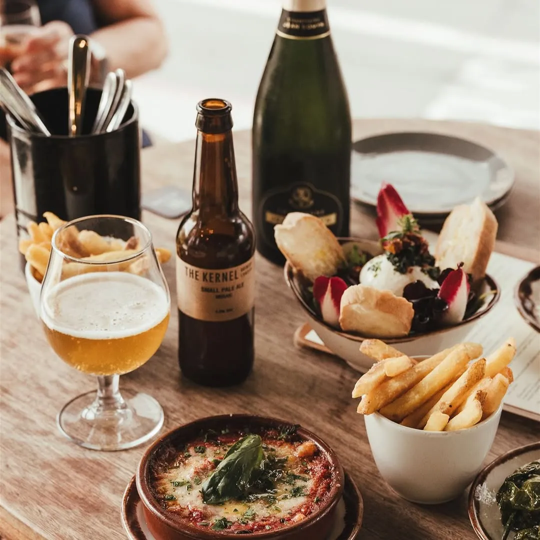 A well-lit and sharp photograph showcasing a variety of food and drinks on a wooden table, including a glass of beer, a beer bottle, a champagne bottle, a bowl of fries, a baked dish, and a salad. The composition is appealing, highlighting the meal in a restaurant setting.