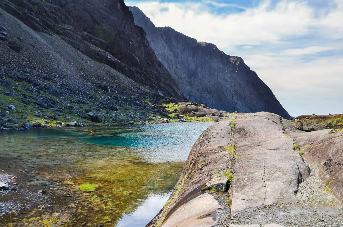 A natural landscape featuring a serene, clear blue-green mountain lake with large rock formations in the foreground and majestic dark mountains towering in the background under a partly cloudy sky.