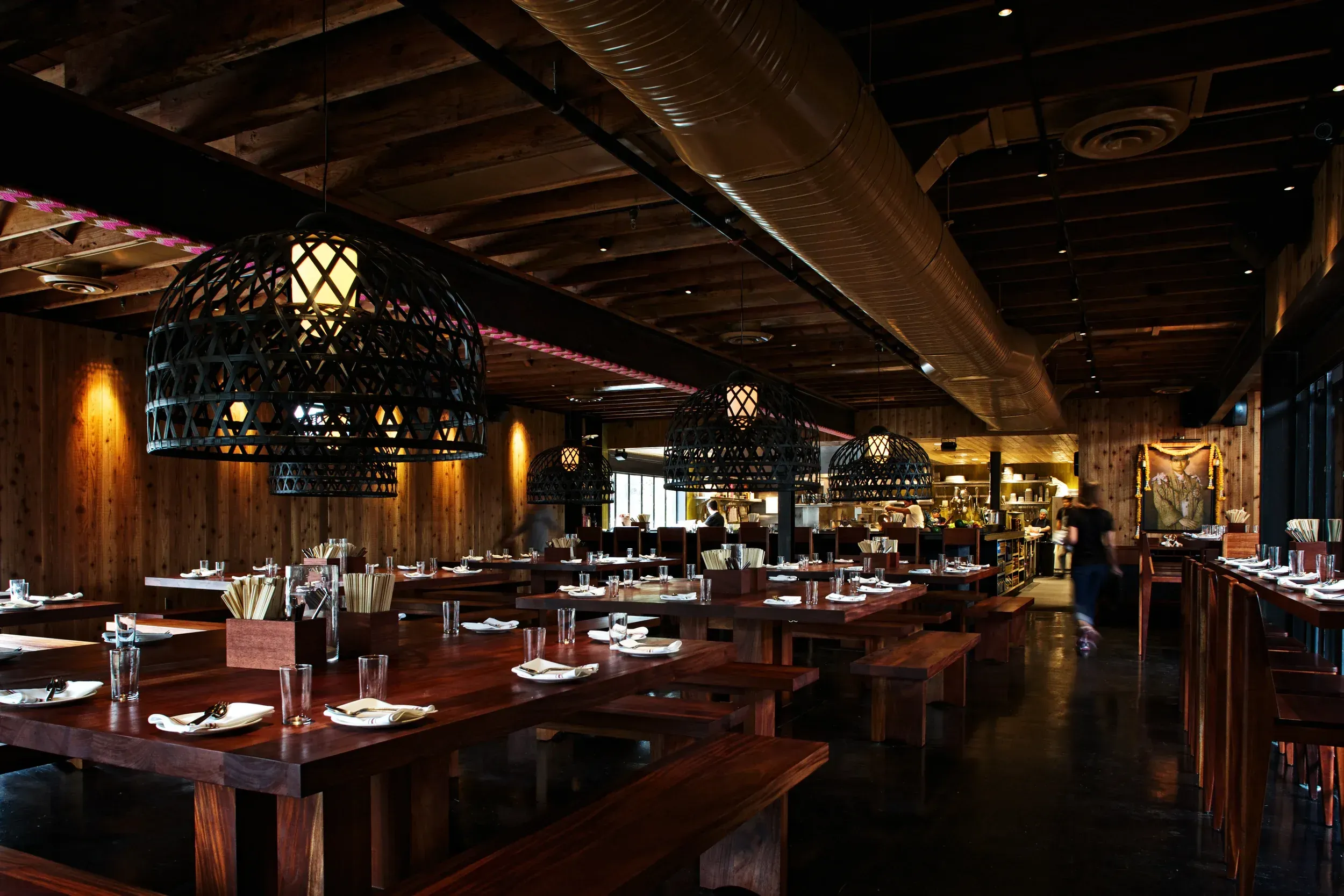 An inviting view of a restaurant's interior featuring dark wooden communal tables and large, intricately designed hanging lamps, creating a warm and atmospheric ambiance. The composition effectively showcases the dining space, highlighting its unique design elements.