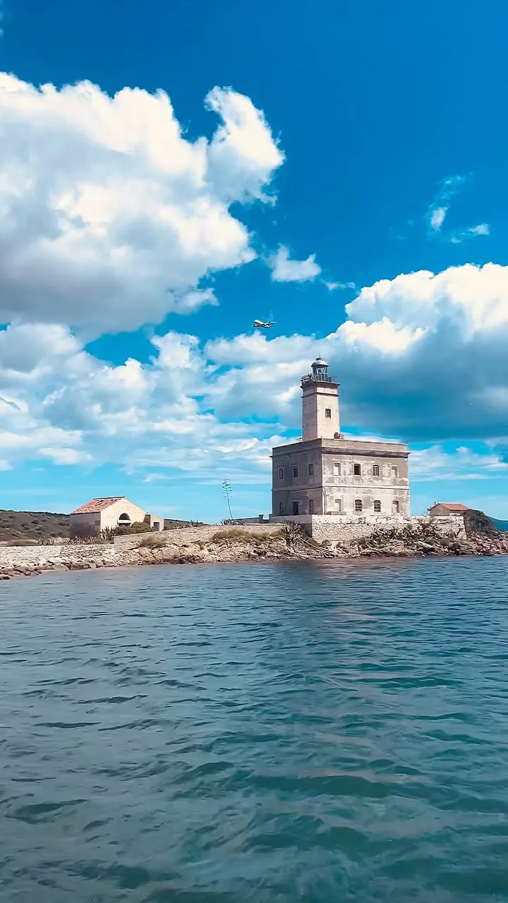 An outdoor photograph featuring a historic lighthouse situated on a rocky island, surrounded by blue water under a bright sky with white clouds. A small airplane is visible in the upper part of the sky.