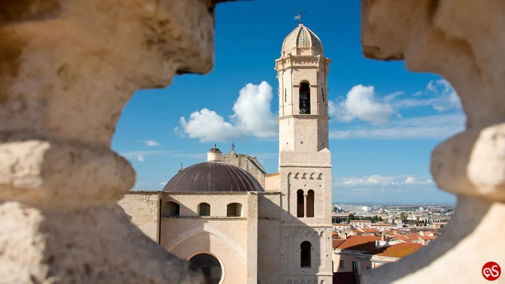 An editorial-style image showcasing a historic church or cathedral with a prominent bell tower and dome, framed by architectural elements in the foreground. The scene is set under a clear blue sky with white clouds, with a distant city skyline visible in the background.