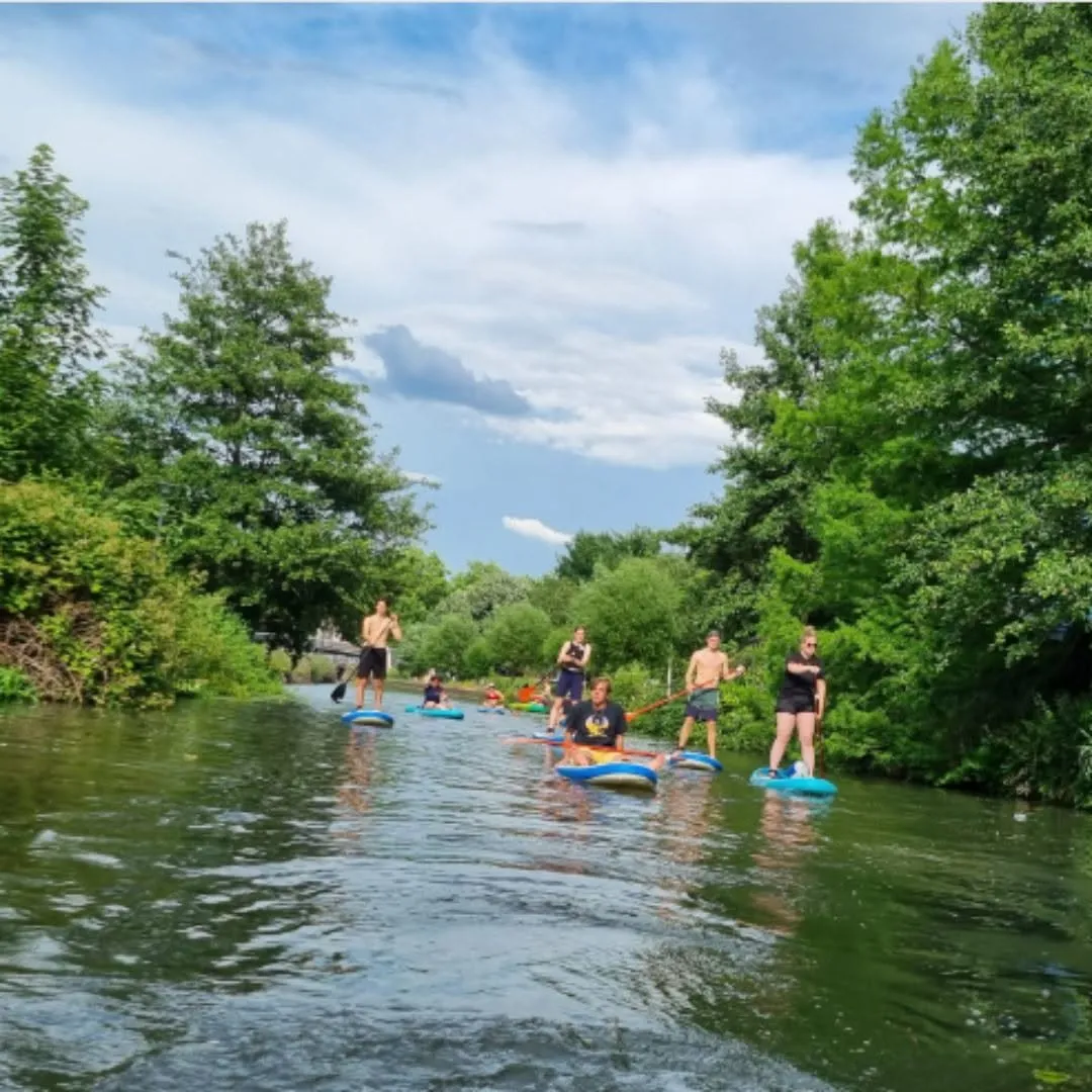 A group of people are enjoying paddleboarding on a calm river, surrounded by lush green trees under a bright, cloudy sky.