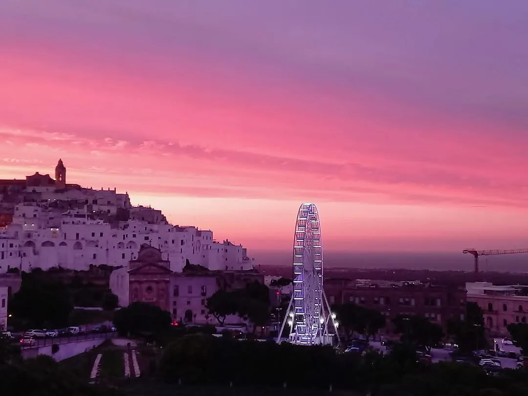 The image captures a picturesque cityscape at sunset, featuring white buildings and a illuminated Ferris wheel against a vibrant pink and purple sky. The overall scene is aesthetically pleasing and visually striking.