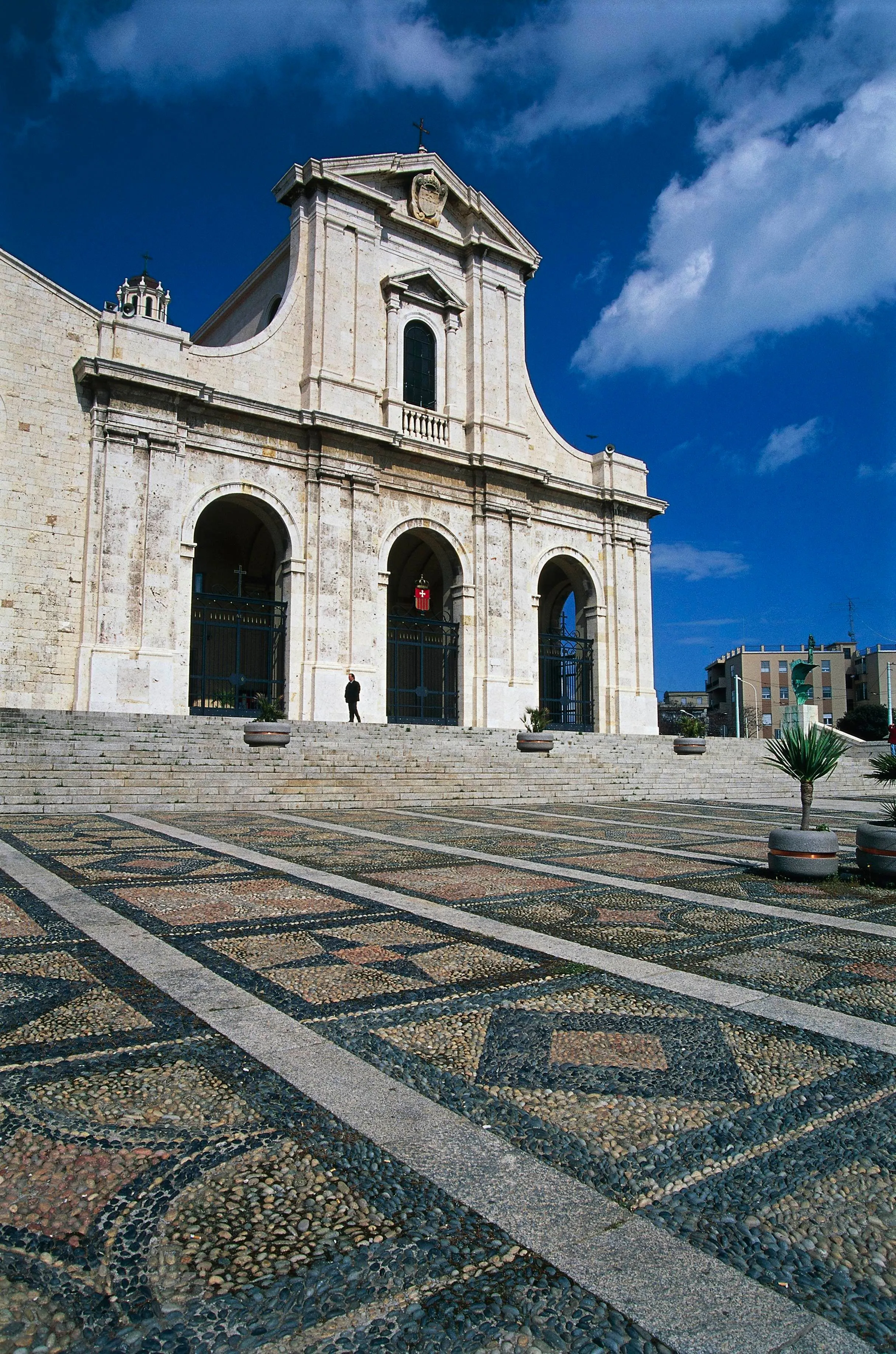 An exterior shot of a grand stone church with a wide plaza in the foreground featuring intricate patterned paving. The sky is bright blue with scattered clouds. A lone figure is visible on the church steps, providing a sense of scale.