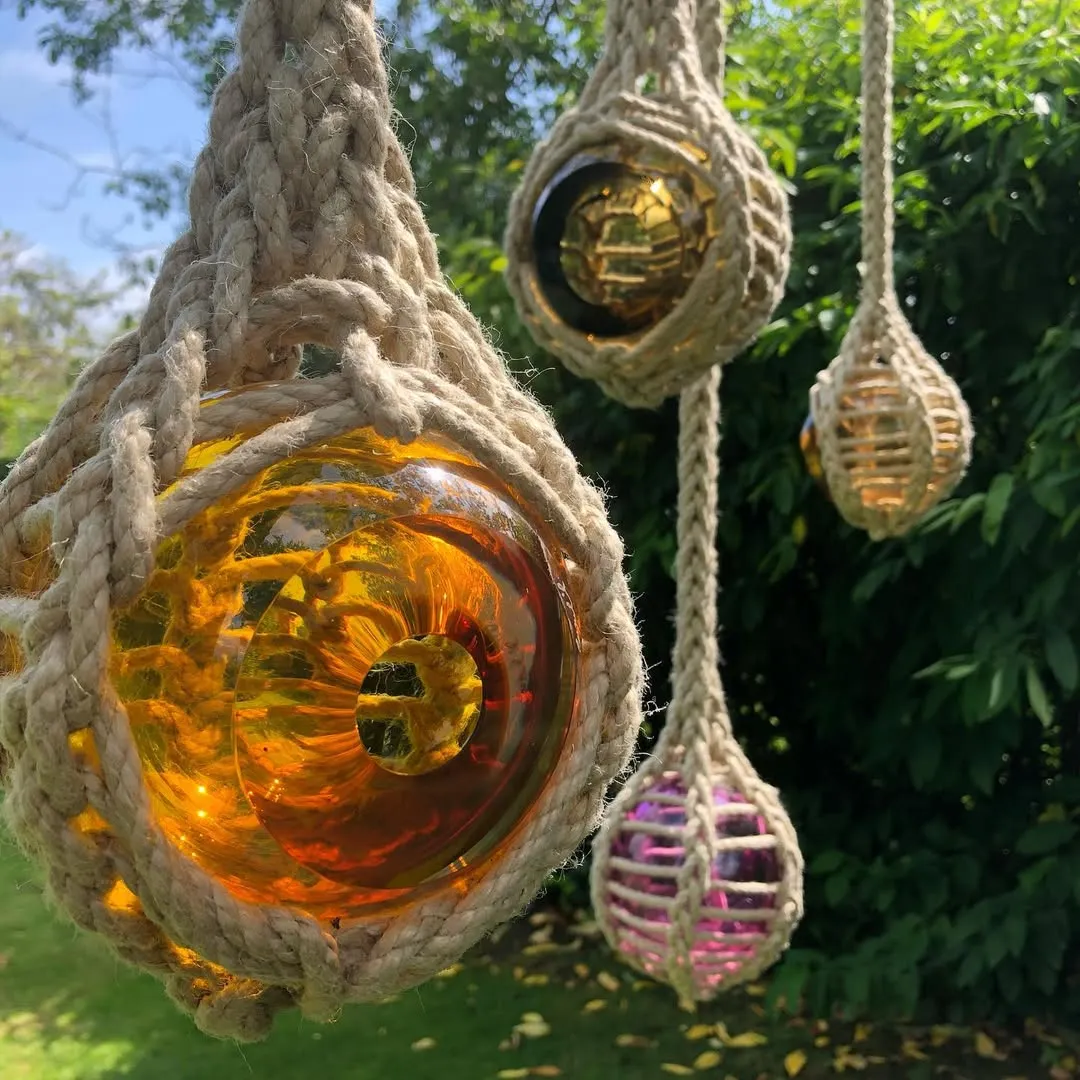 A close-up shot of several decorative glass floats wrapped in thick rope, hanging outdoors against a lush green, blurred background. The main float in the foreground is a vibrant amber/yellow, while others in the background are also amber and one appears magenta. Natural light illuminates the objects, highlighting their texture and color.