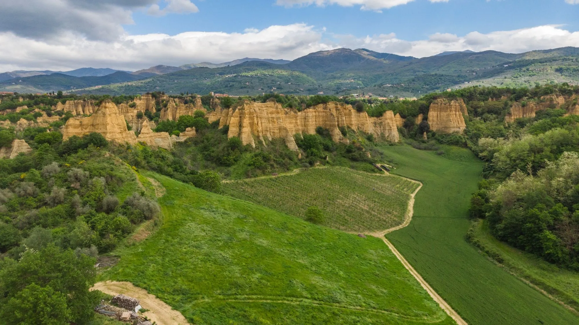 An expansive aerial view showcasing a diverse natural landscape, featuring striking sandy badlands, lush green agricultural fields, winding paths, and distant verdant mountains under a dynamic sky with white clouds.