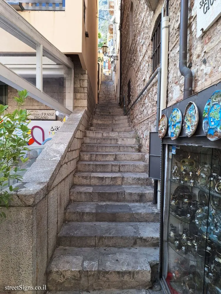 A narrow, stone-paved alleyway with a long flight of stone stairs leading uphill between old buildings. A shop display with ceramic plates and other items is visible on the right, and a green plant adds a touch of nature on the left. The scene captures the essence of an old European town street.