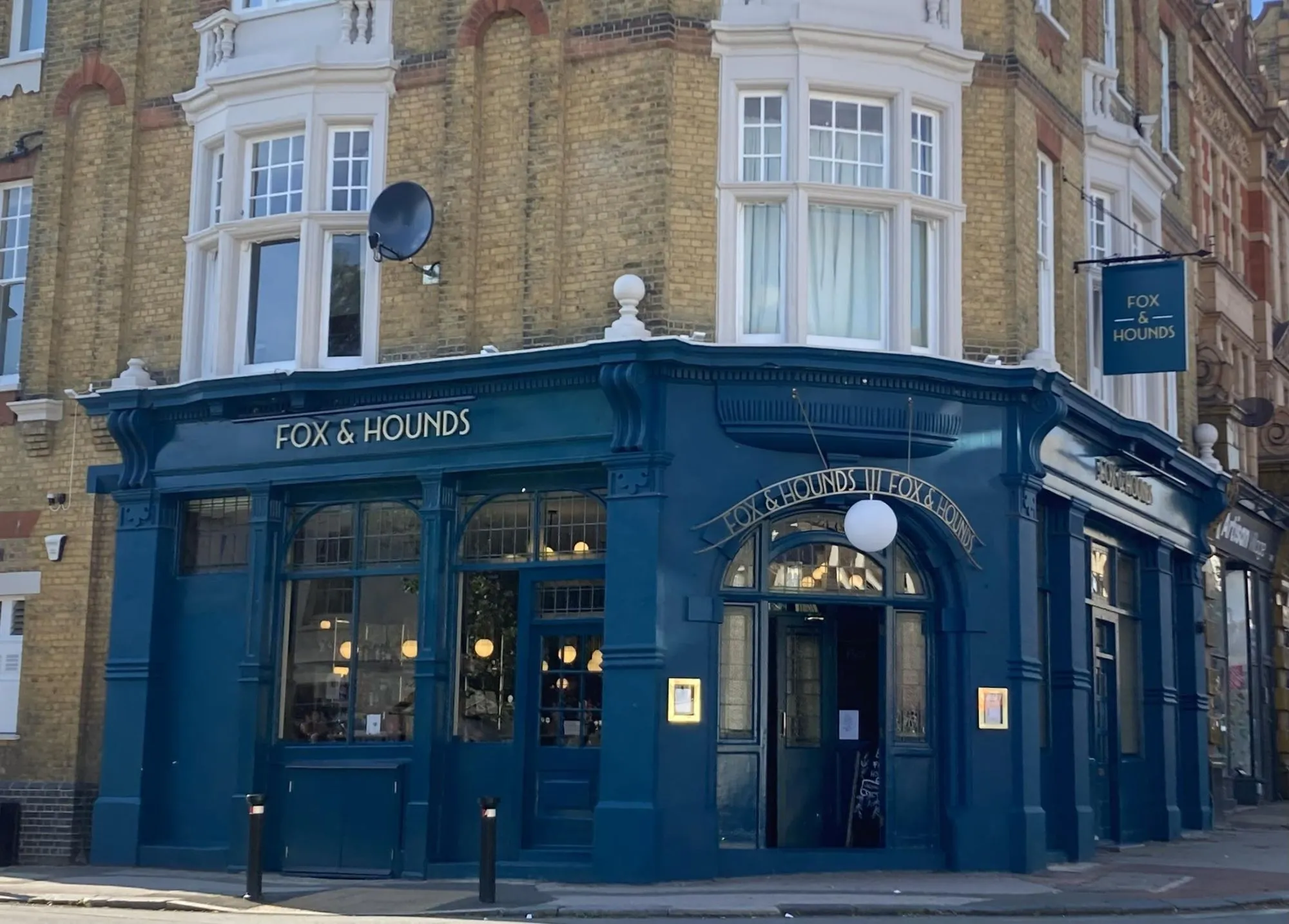 A well-composed and sharp photograph of a traditional pub exterior, 'Fox & Hounds', featuring its distinctive blue facade and signage. The natural lighting is good, contributing to accurate colors and high photo-realism. All key elements, including the pub name and entrance, are in focus, making for a clear and engaging image.
