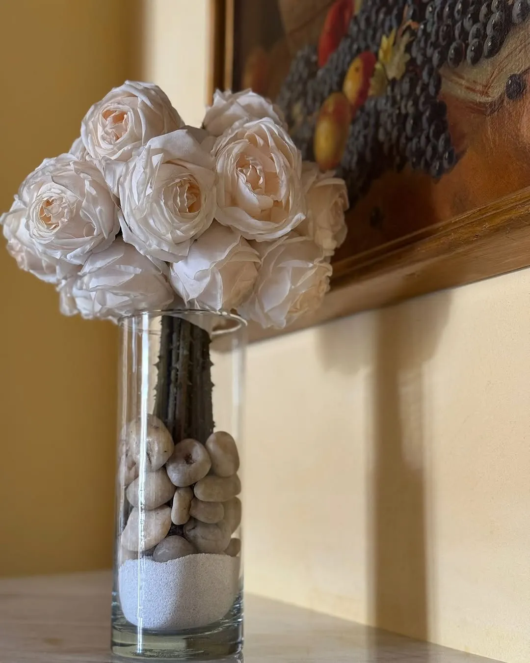 A still life image featuring a bouquet of white roses in a clear glass vase, filled with decorative rocks and sand. The vase is placed on a surface with a framed painting in the background, against a yellow wall.
