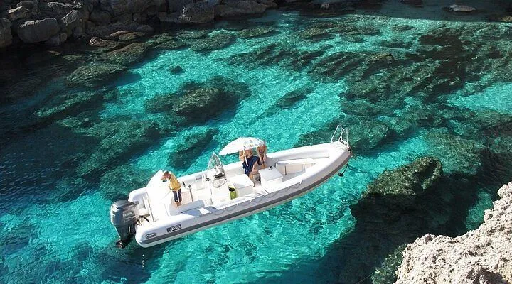 An aerial view of a white boat with people on board, floating on incredibly clear, turquoise-blue water, revealing the rocky seafloor beneath. The scene suggests a tranquil, sunny day at a beautiful marine location.